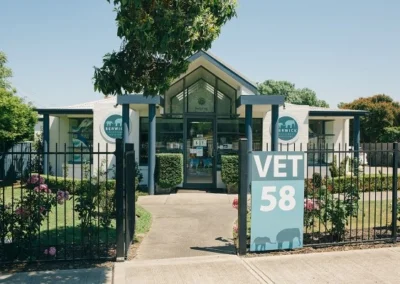 Front view of Berwick Veterinary Clinic with a sign reading “VET 58,” glass entrance, manicured garden, and black fence on a sunny day.