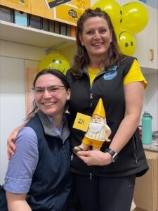 Two Berwick Veterinary Hospital staff members smiling together during an R U OK Day event, holding a small gnome and surrounded by yellow balloons and decorations