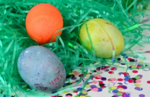 Three colourful Easter eggs resting in bright green decorative grass beside scattered confetti
