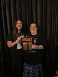 Mandy Villinger award presentation photo showing two women holding an award plaque in front of a black curtain backdrop