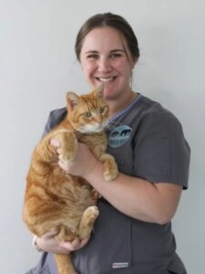 Nurse Kelsey smiling while holding an orange tabby cat in her arms against a plain background.
