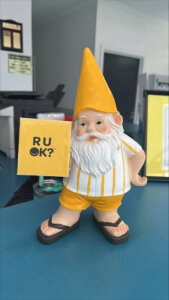 Small garden gnome figurine holding a yellow “R U OK?” sign displayed on a veterinary clinic reception desk during an R U OK Day awareness event