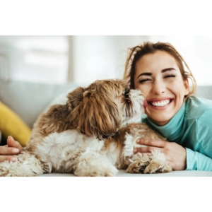 Woman smiling while a small fluffy dog affectionately licks her cheek as they lie together on a couch at home